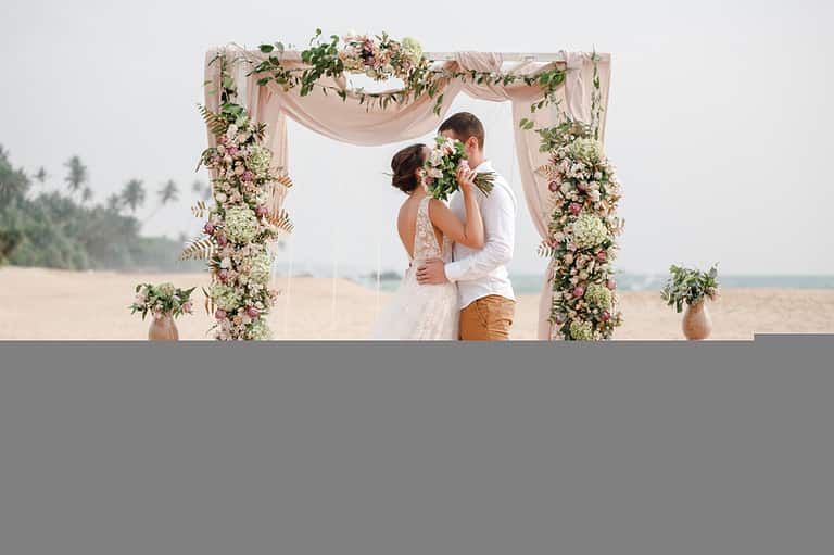 bride and groom standing on beach under flower arbor