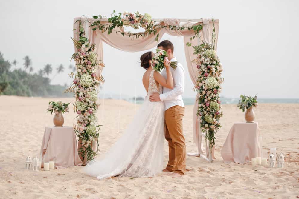 bride and groom standing on beach under flower arbor