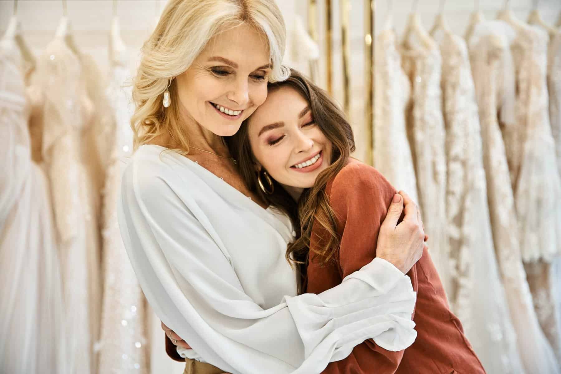 middle-aged woman hugging younger woman in front of a rack of wedding dresses