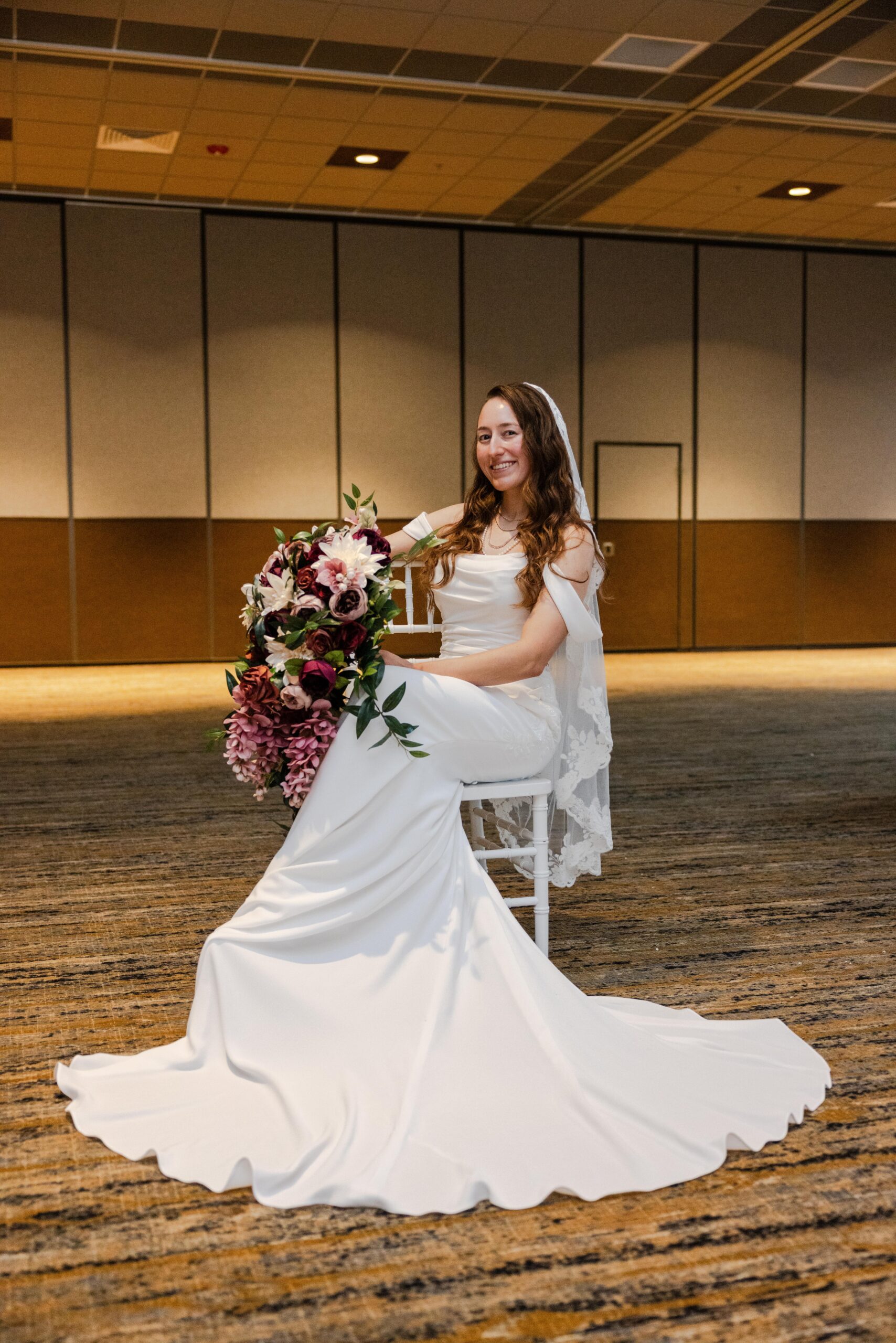 Savvy bride Mikayla sitting on a white chair wearing her wedding dress while holding a bouquet of burgundies color flowers.