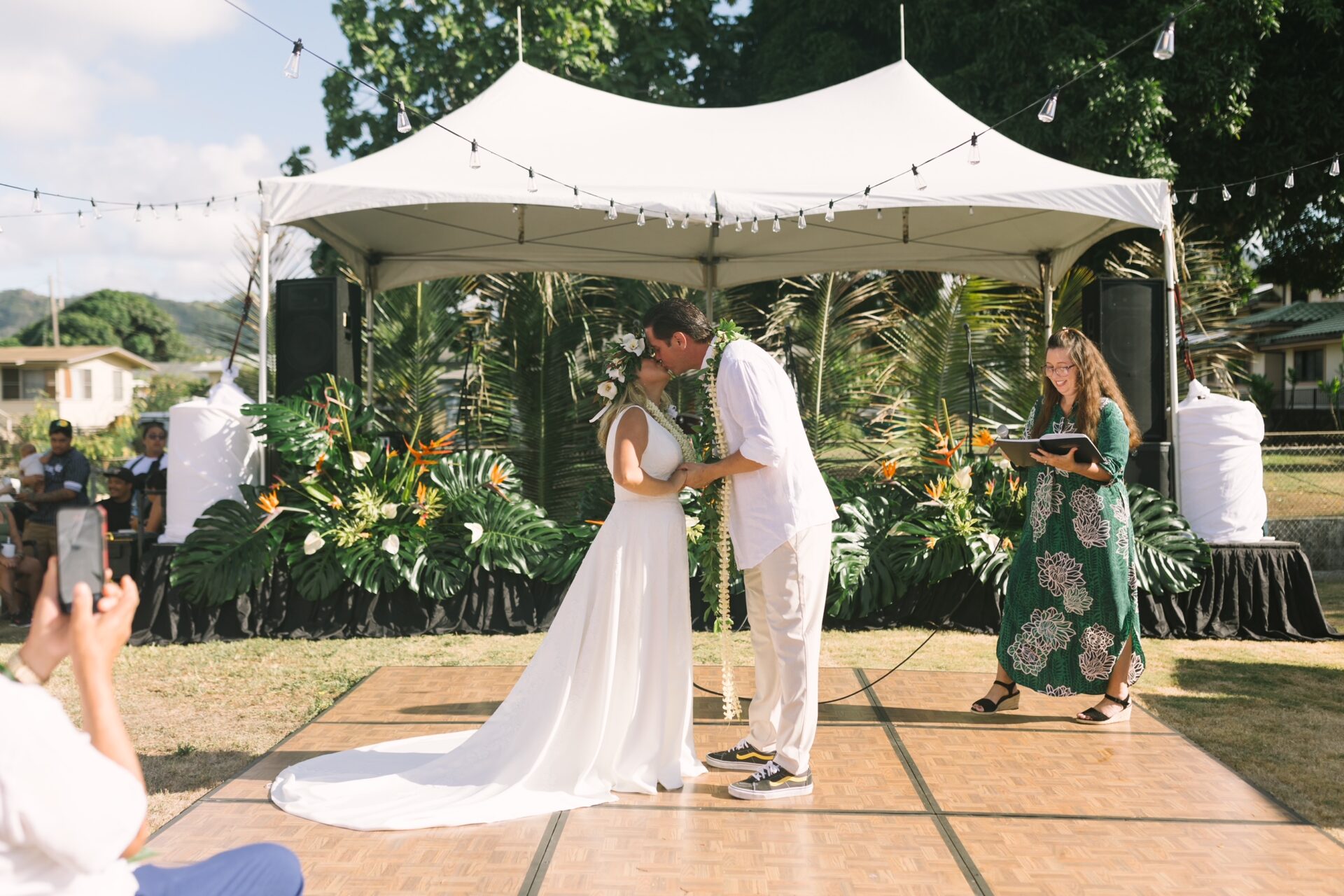 The couple first kiss after the wedding ceremony. The stunning background complete with an airy white tent, warm string lights, and tropical florals arranged in lush, textural displays. Anthuriums, monstera leaves, and island greenery added the perfect touch of natural sophistication.