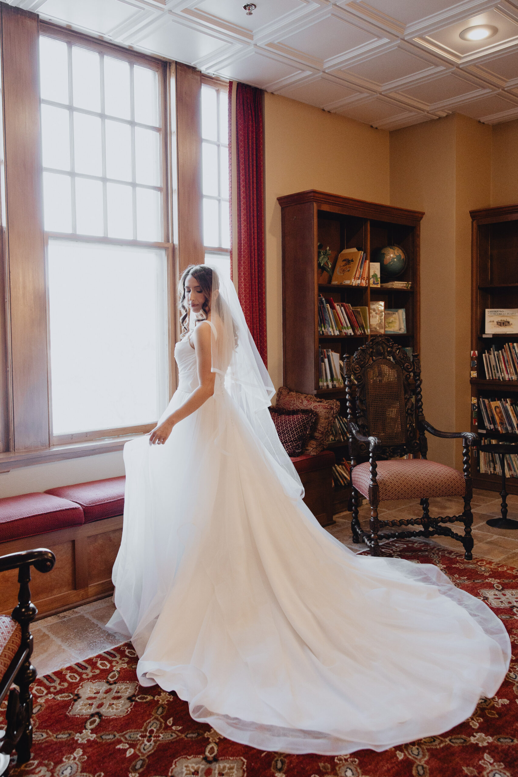 woman standing near a wide window wearing a white A-line  wedding dress featuring a structured bodice and layers of ethereal, frothing tulle paired with veil to complete her look.