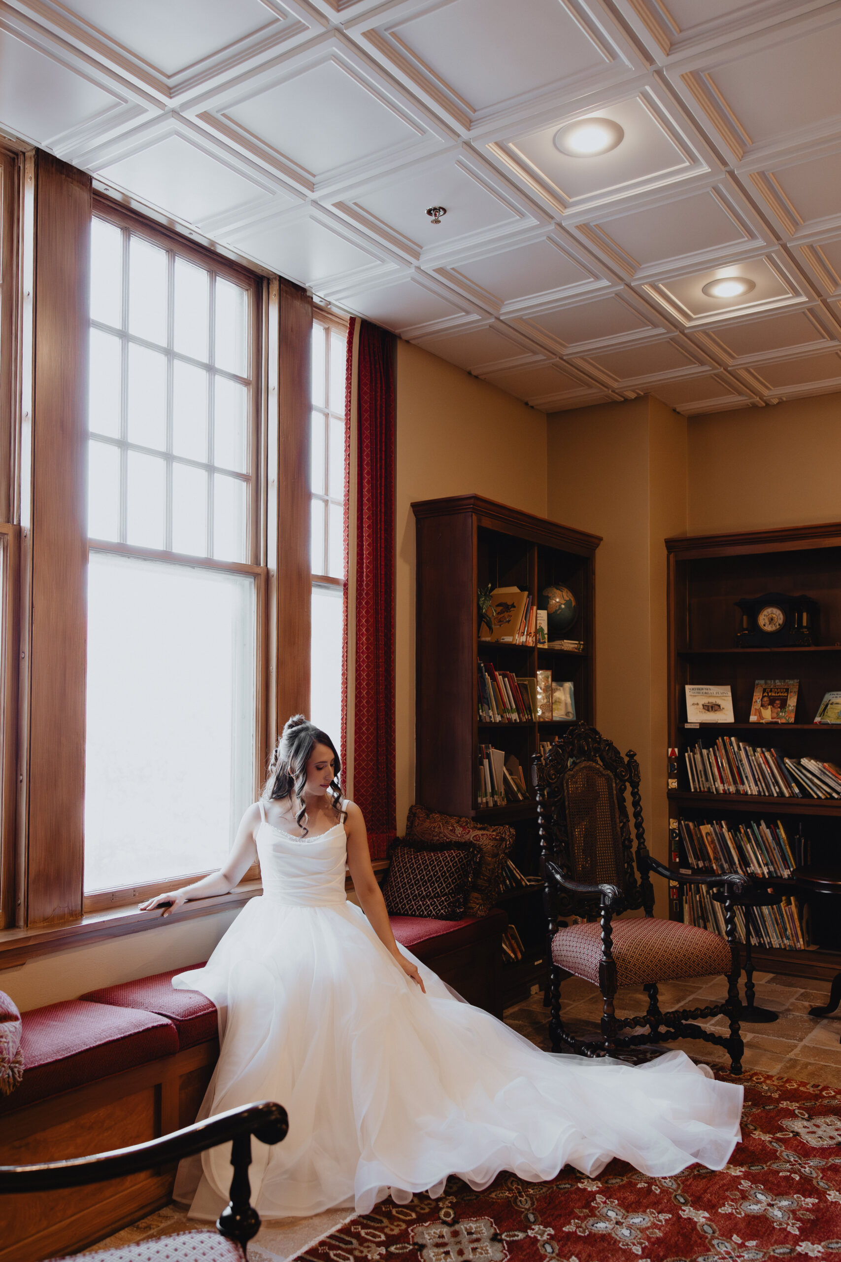 woman comfortably setting near a wide window wearing a white A-line  wedding dress featuring a structured bodice and layers of ethereal, frothing tulle.