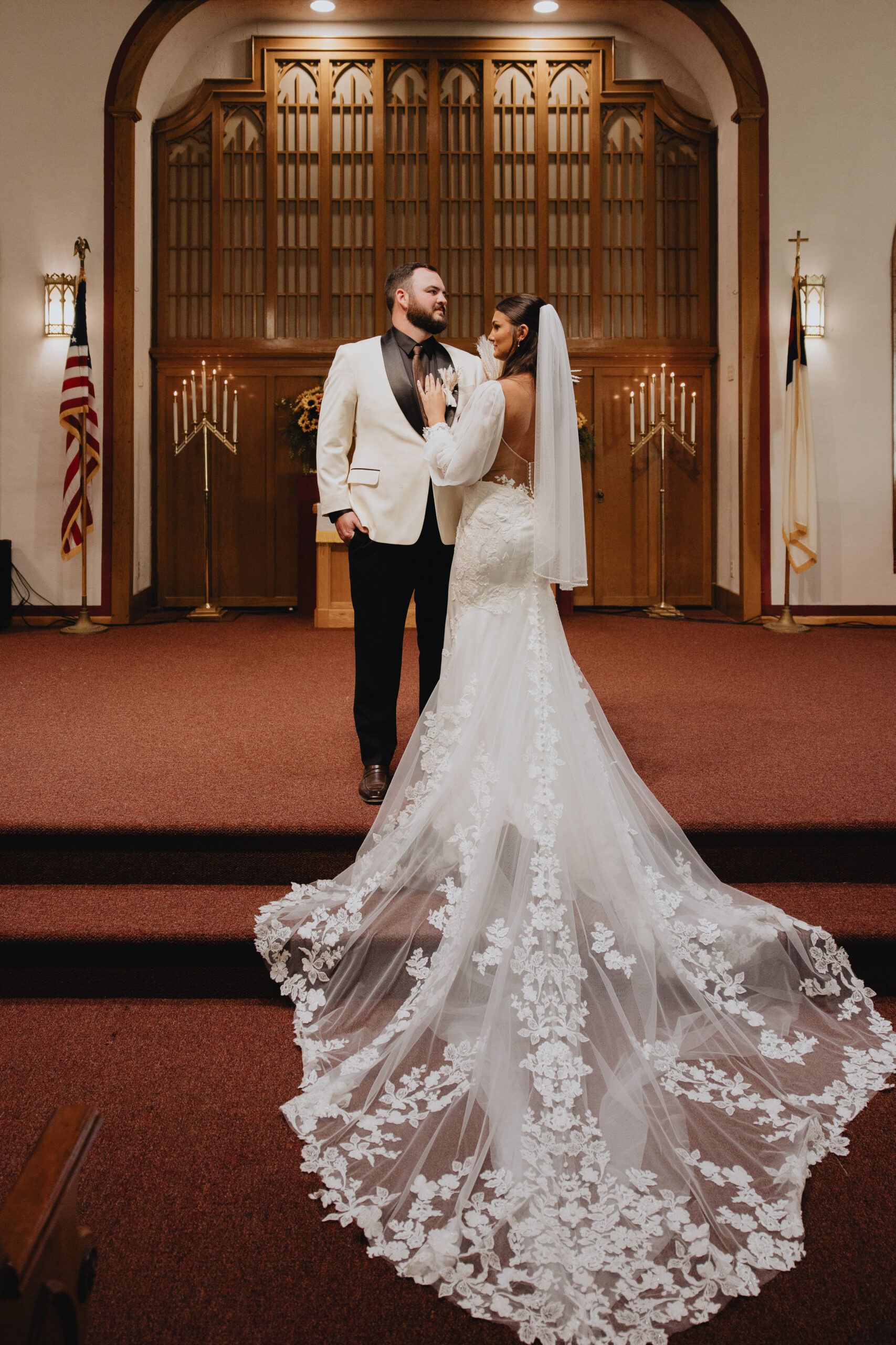 Kelsey the bride wearing an exquisite lace detailing, sheer statement sleeves that added a whimsical boho touch, and a dramatic train with intricate floral appliqués paired perfectly with Colby stood out in a stylish white tuxedo jacket with black lapels.
