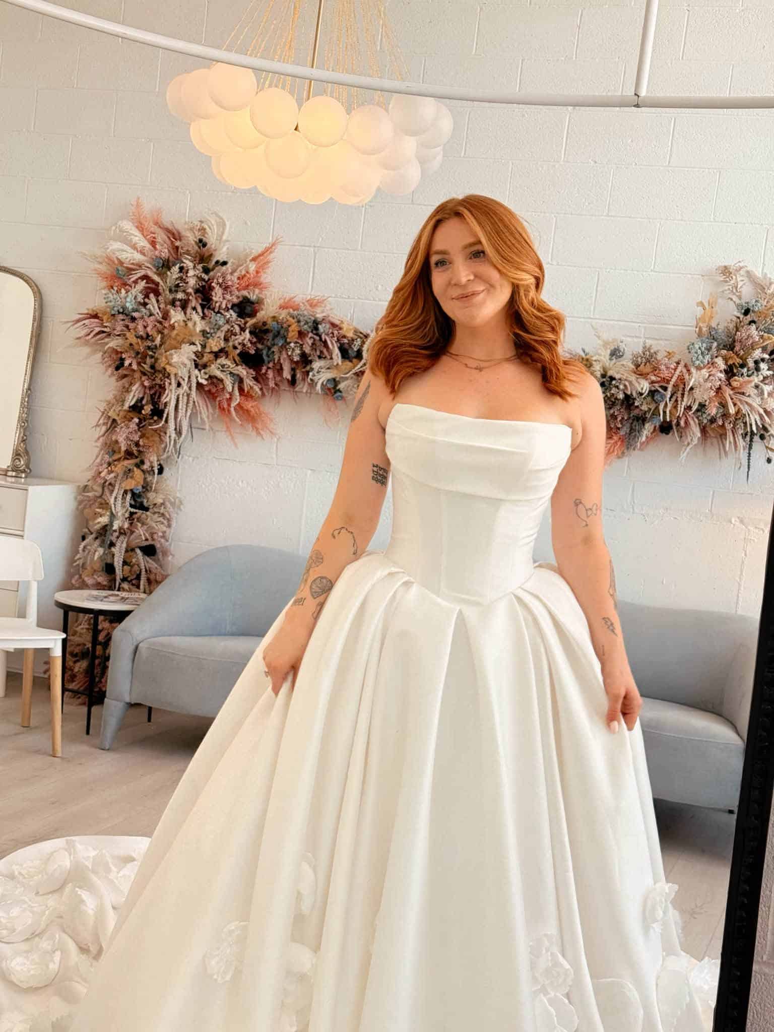 Bride wearing a strapless satin Basque waist ball gown wedding dress with a structured corset bodice, elegant draped neckline, and full skirt featuring delicate 3D floral appliqués, photographed inside Savvy Bridal boutique.