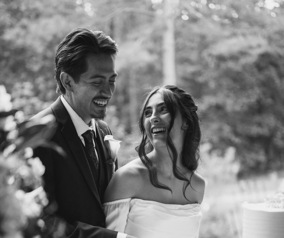 Savvy Bridal bride Brenda and groom Saul sharing a joyful moment outdoors, with the bride wearing an elegant off-the-shoulder satin wedding dress and smiling at her partner on their wedding day.