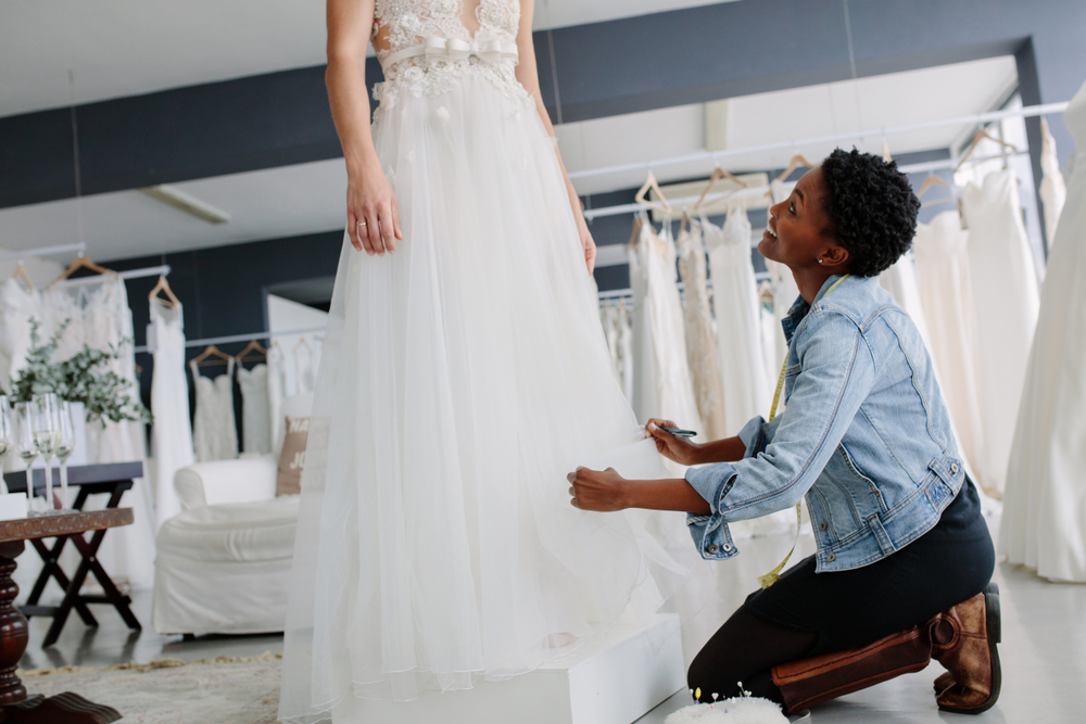 Woman making adjustment to bridal gown in wedding fashion store. Bride in her wedding dress with female dress designer making final adjustments on dress and smiling.
