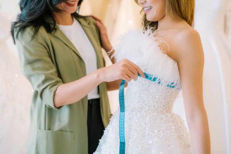 Asian bride is smiling while choosing wedding dress in modern wedding salon, Attractive designer girl using tape meter fitting on wedding dress at wedding studio
