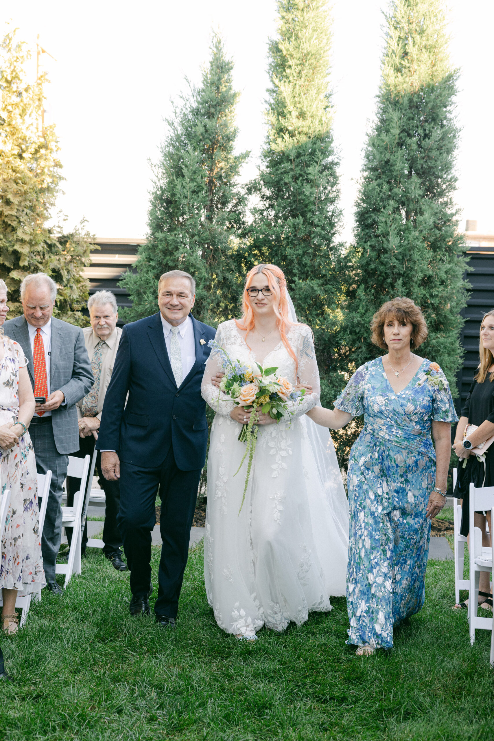 Caitlyn walking down the aisle in a lace wedding gown with family during her outdoor wedding ceremony.