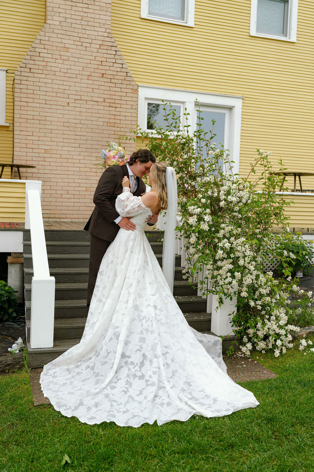 Bride and groom kissing outdoors beside a yellow wedding venue.