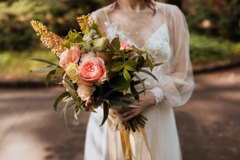 Bride holding a romantic bouquet of roses and greenery in soft natural light. Elegant wedding dress with delicate details, outdoor forest setting, intimate bridal portrait