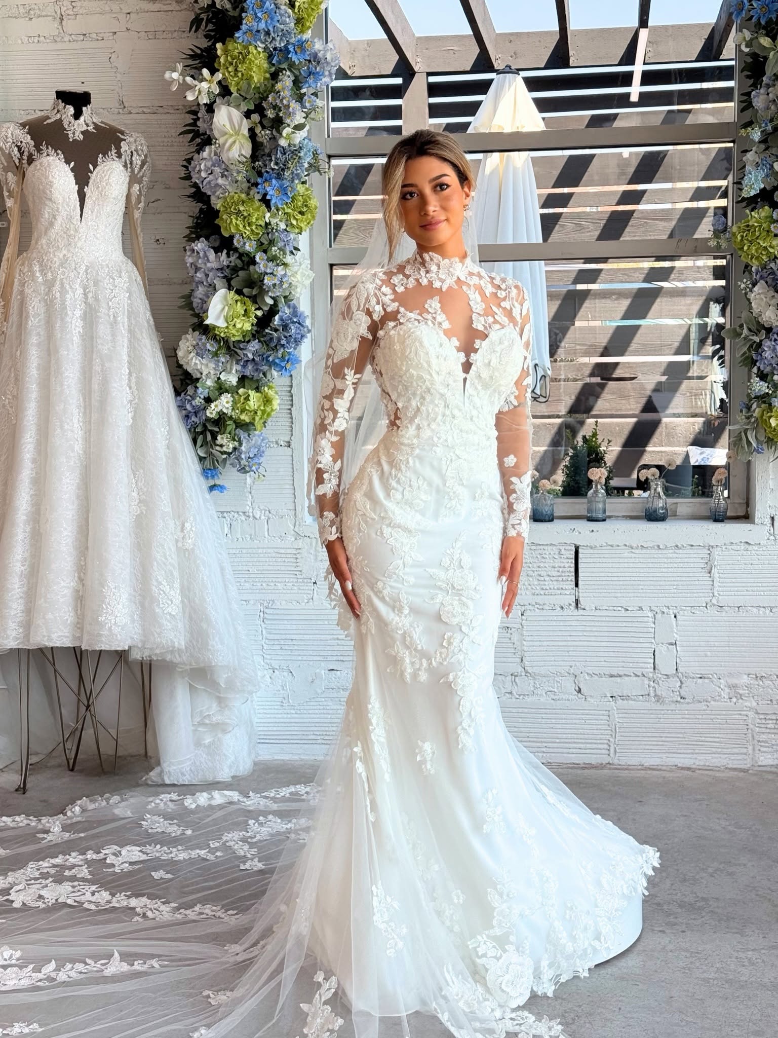Bride wearing a fitted floral lace wedding dress with sheer long sleeves, a high neckline, and a flowing train at Savvy Bridal.