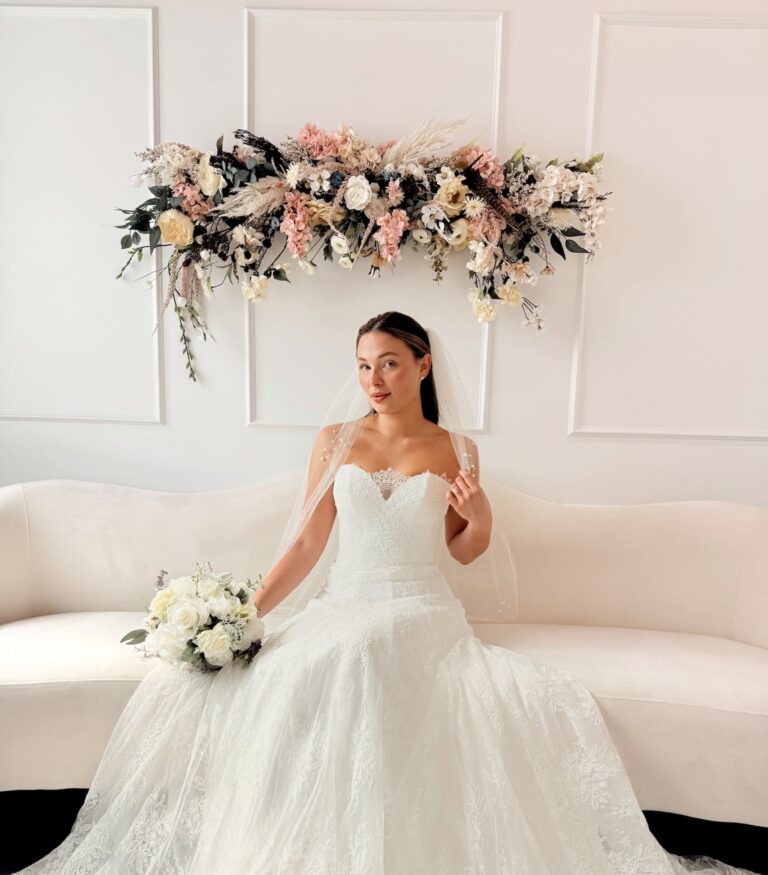 Bride seated in a romantic lace wedding gown with a flowing skirt and veil, holding a white rose bouquet.