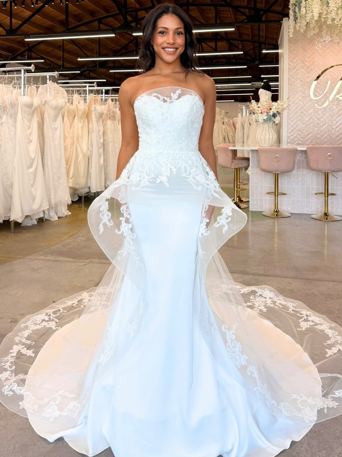 Model wearing a strapless wedding gown with floral lace details and a dramatic sheer overskirt at Savvy Bridal.