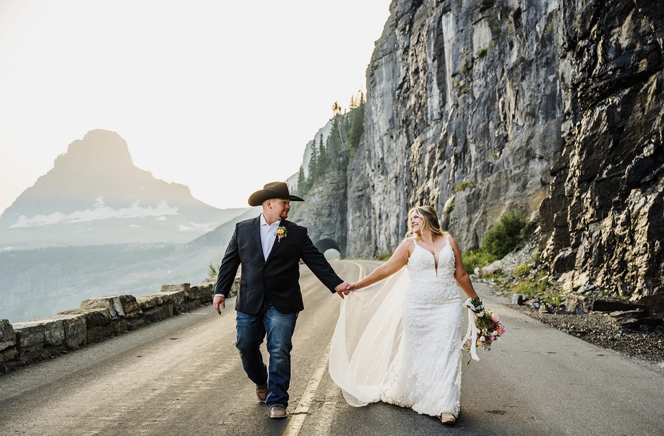 Annabelle in her romantic floral wedding dress holding hands with Zack along a scenic mountain road in Glacier National Park.