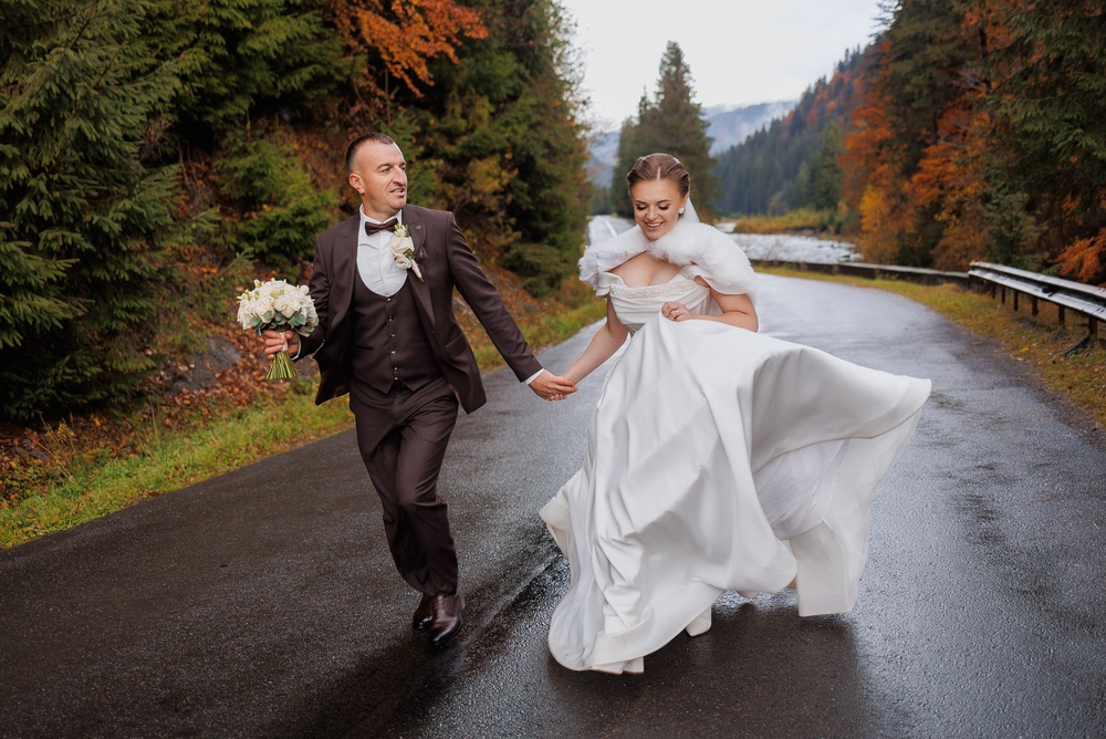 A joyful bride and groom run hand in hand along a wet road surrounded by colorful autumn trees. They are filled with love and excitement on their wedding day in a picturesque landscape.